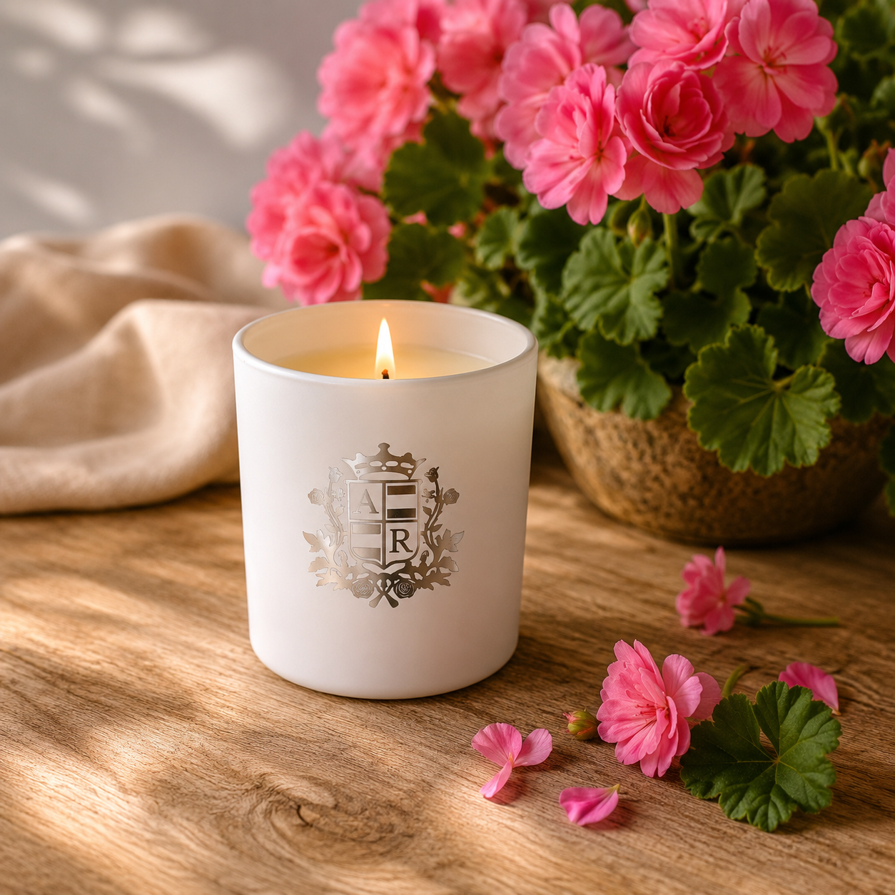A white ceramic candle jar with a silver monogram crest sits on a wooden table, lit with a small flame. Pink flowers and green leaves in a pot are blurred in the background, along with a beige cloth and scattered flower petals.