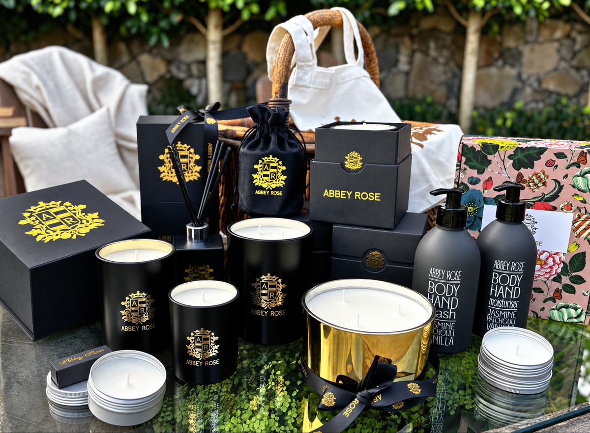 Collection of Abbey Rose products including candles, body wash, and other items on a reflective surface with greenery in the background.