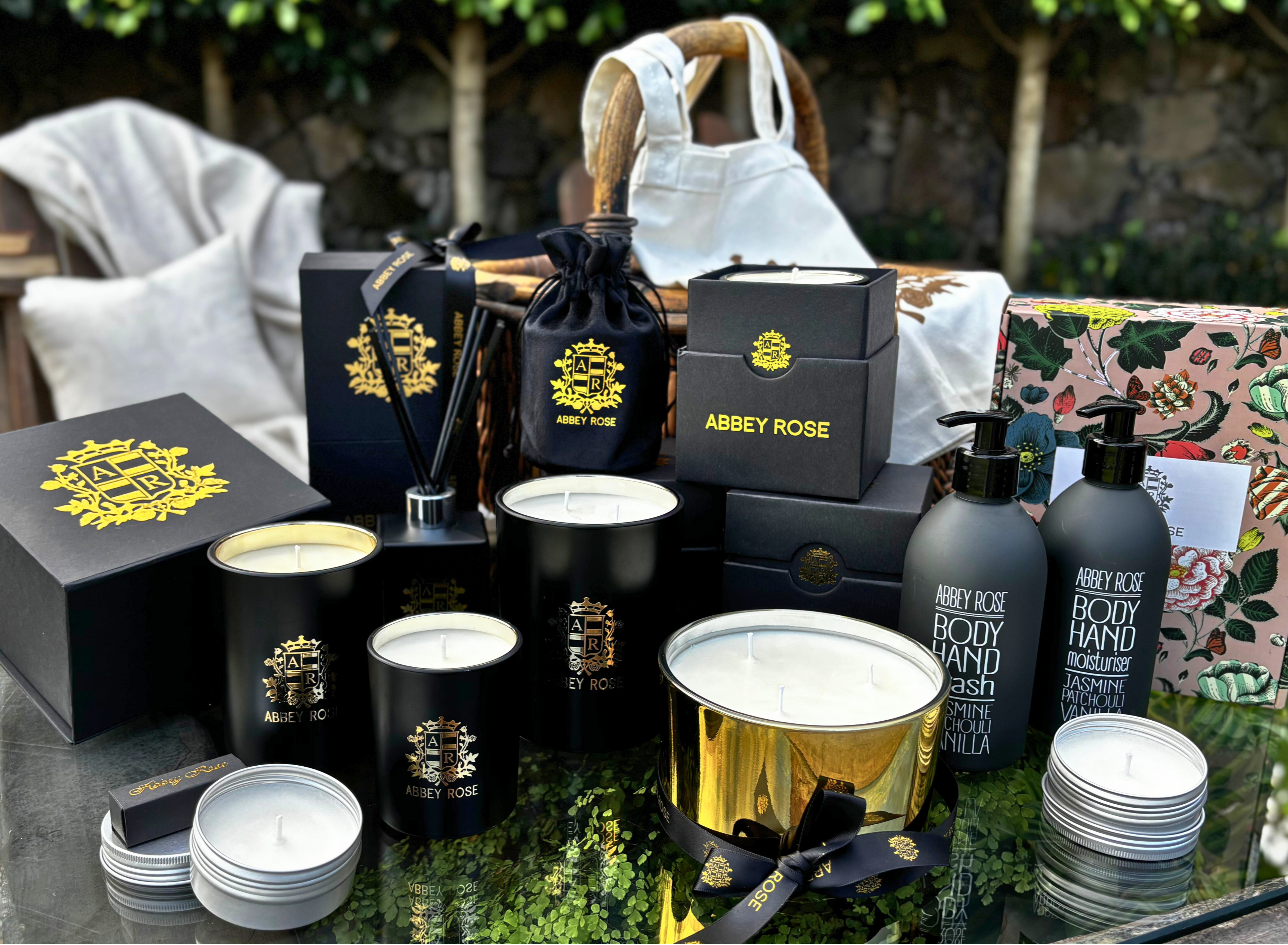 Collection of Abbey Rose products including candles, body wash, and other items on a reflective surface with greenery in the background.