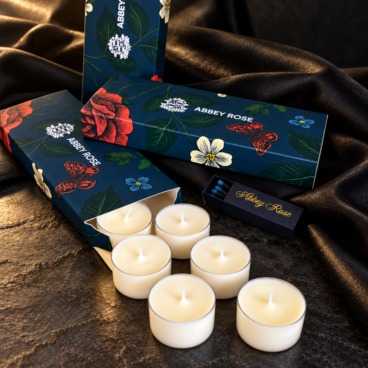 Abbey Rose Discovery Set tealight candles beside navy floral gift boxes and a branded matchbox on dark stone with warm gold lighting.